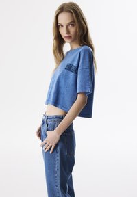 Blaues, kurzes T-Shirt aus weichem Material mit Rundhalsausschnitt und kurzen Ärmeln; hat schwarze Textakzente auf der Vorderseite und wird mit Jeans aus Denim kombiniert.