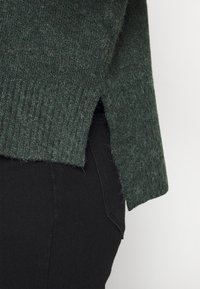 Ciemnozielony sweter z teksturowanym splotem, bocznymi rozcięciami i ściągaczem u dołu, zestawiony z czarnym dżinsowym materiałem. Bez widocznych logo ani wzorów.