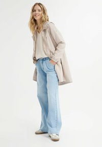 Cappotto beige con cappuccio, maglione a righe grigio chiaro e jeans a gamba larga blu chiaro. Sneakers beige con accenti bianchi. Sfondo bianco semplice.