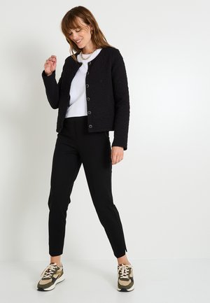 Veste matelassée noire avec col rond, boutons-pression, portée sur un t-shirt blanc et un pantalon noir ajusté, associée à des baskets multicolores.