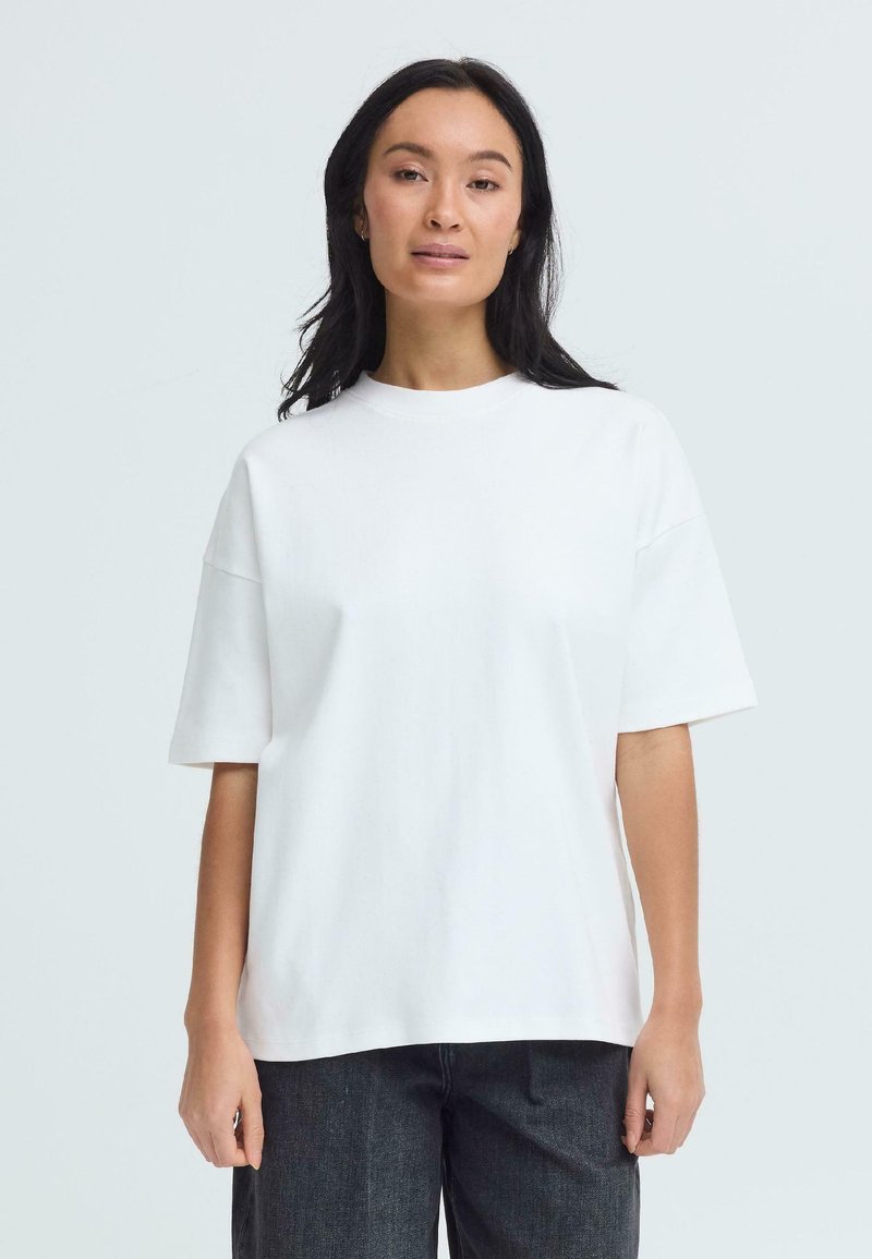Femme aux longs cheveux noirs portant un T-shirt blanc uni à manches courtes et ample, ainsi qu'un pantalon sombre, debout devant un fond clair.
