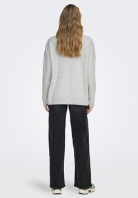 Jasnoszary sweter z dzianiny o luźnym kroju i ściągaczu u dołu, zestawiony z ciemnymi szerokimi dżinsami i sneakersami. Teksturowany, casualowy wygląd.