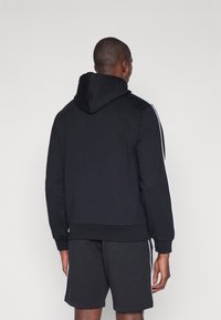 Sweatshirt preto com capuz, feito de uma mistura de algodão, com riscas brancas laterais, punhos canelados e capuz com cordão. Usado com calções combinando.