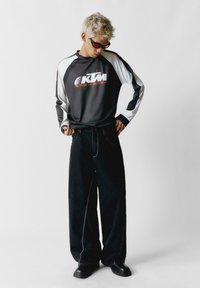 Čierne veľké nohavice s bielym prešívaním a uvoľneným strihom. Dlhý rukáv v šedej a bielej farbe s oranžovým brandingom KTM. Čierne slnečné okuliare.