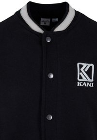 Veste de style varsity noire avec un col côtelé blanc, des boutons-pression argentés et un logo "KANI" brodé sur la poitrine gauche.