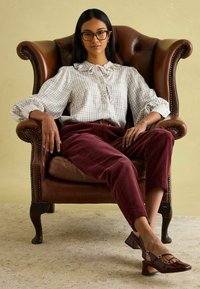 Karierte Bluse mit Rüschenausschnitt, burgunderfarbene Cordhosen und braune Lackpumps. Frau, die in einem braunen Lederarmstuhl vor einem hellen Hintergrund sitzt.