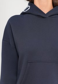 Suur lähedalt vaade tumesinisele kapuutsiga džemprile, millel on eesmine tasku, kanda inimene, kellel on pikad tumedad juuksed, ühevärvilise heleda tausta ees.