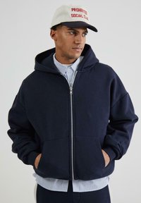Marineblå zip-up hoodie i blødt materiale med store frontlommer, båret over en stribet skjorte med knapper. Akkompaneret af en hvid og marineblå cap.