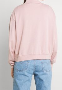 Sudadera rosa con cuello alto y hombros caídos, que presenta una textura de tela suave y dobladillo acanalado, combinada con jeans de color azul claro.