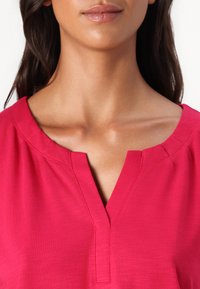 Rosa Kurzarmshirt mit V-Ausschnitt, das feine Textur- und Nähdetails am Kragen aufweist. Leichter Stoff, lässiges Design.