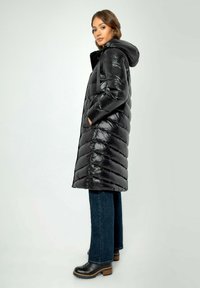 Langer schwarzer Puffercoat mit einer glänzenden, gesteppten Textur, ausgestattet mit einer Kapuze und seitlichen Taschen, kombiniert mit dunklen Jeans und klobigen schwarzen Stiefeln.