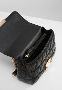 Černá kožená taška s prošíváním a zlatým zapínáním. Má vnitřní kapsu na zip a řetízkový popruh. Hladká textura s pevnou konstrukcí.