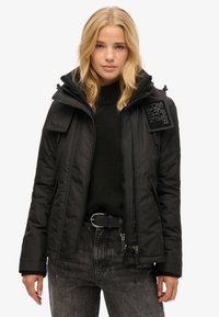 Veste noire isolée avec fermeture éclair à l'avant, capuche amovible et deux poches zippées. Présente des poignets côtelés et une étiquette logotée sur l'épaule.