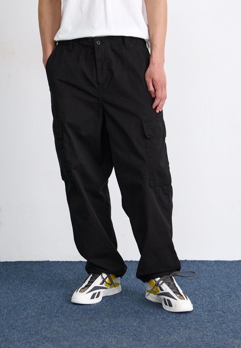 Pantaloni cargo negri cu multiple buzunare, realizați dintr-un material durabil. Croi lejer cu închidere prin buton, asortați cu adidași albi și gri.