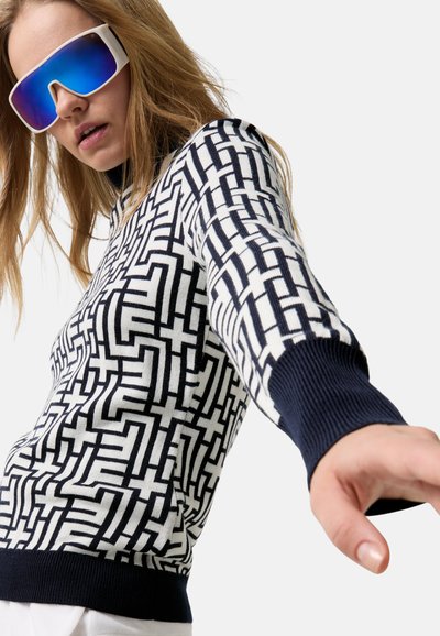 Kobieta z długimi włosami, nosząca duże niebieskie lustrzane okulary przeciwsłoneczne i sweter w czarno-białe geometryczne wzory, sięgająca do przodu.