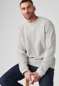 Light gray ribbed fabric sweatshirt with long sleeves and a crew neckline, paired with dark blue jeans, seated on a wooden stool.