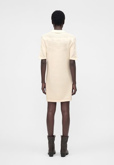 Γυναίκα με κρεμ, κοντομάνικο, φόρεμα μήκους μέχρι το γόνατο με υφή κεντήματος στην πλάτη και καφέ μποτάκια, στέκεται μπροστά σε λευκό φόντο.