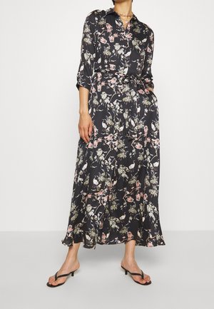 Femme portant une robe longue à manches longues noire à motifs floraux avec des boutons et une ceinture à la taille, ainsi que des sandales à talons noires, debout devant un fond uni.