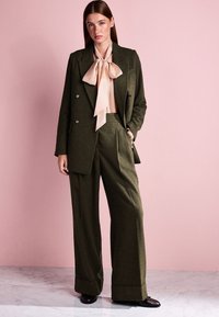 Olive green tailored suit with wide leg pants and double-breasted blazer. Paired with a light pink blouse featuring a large bow tie.