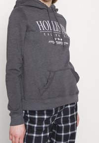 Grå huvtröja med framficka och vittryckt "Hollister California"-logotyp. Har ribbad muddar och nederkant. Kombineras med svarta rutiga byxor.
