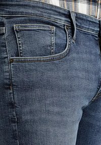 Zbliżenie detali kieszeni niebieskich dżinsów oraz pasa, z widocznym przeszyciem i częścią koszuli w kratę włożoną w spodnie.