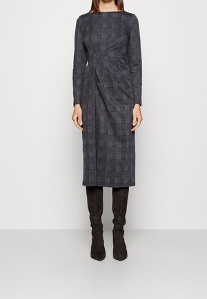 Femme portant une robe grise à carreaux manches longues, longueur genou, avec des plis asymétriques à l'avant, associée à des bottes noires montant jusqu'au genou.