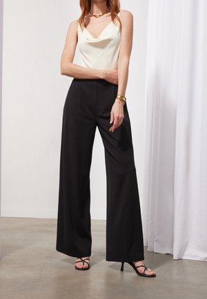 Femme portant un haut blanc sans manches, un pantalon large noir taille haute, des talons noirs à brides, un collier et un bracelet dorés, debout à l'intérieur.
