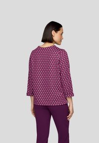 Fuchsia Oberteil mit geometrischem Muster, Dreiviertelärmel, runden Ausschnitt und glattem Stoffe, kombiniert mit lila Hosen.