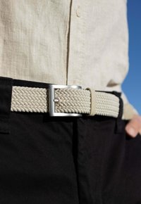 Cintura intrecciata beige con fibbia in metallo argento, indossata sopra pantaloni neri. Presenta una texture a motivo diamante e un dettaglio con asola regolabile.