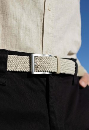 Geweven beige riem met zilveren metalen gesp, gedragen over zwarte broek. Heeft een diamantpatroonstructuur en een verstelbaar lusdetail.