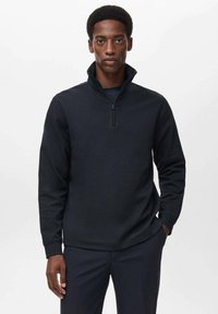 Črna zip-up mikserica z visokim ovratnikom, dolgimi rokavi in teksturirano tkanino. Nosijo jo z temnimi hlačami, oblikovana za udoben fit.