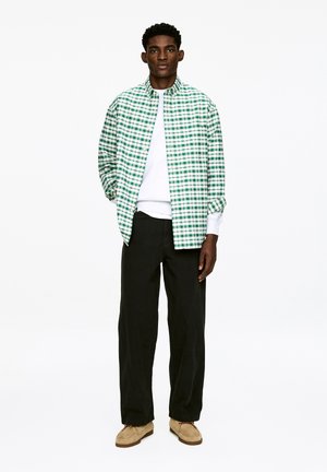 Chemise à carreaux verts et blancs portée sur un pull gris, associée à un pantalon noir et des chaussures beige. La chemise a un col à boutons et des manches retroussées.