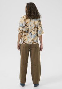 Frau mit lockigem Haar, die ein blau-braunes Batikshirt, braune Hosen und blaue Schuhe trägt, steht im Profil vor einem schlichten weißen Hintergrund.