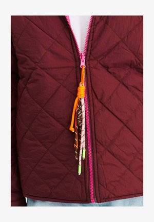 Burgundská prešívaná bunda s ružovým zipsom a farebnými šnúrkami ako doplnkom. Tkanina má mierne lesklú štruktúru s diamantovými vzormi.