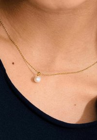 Collier en or avec un petit pendentif en perle, arborant une chaîne délicate et une texture lisse, porté sur un cou nu avec un haut sombre.