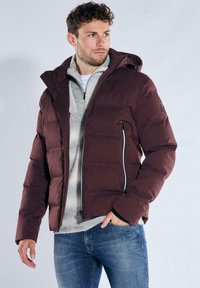 Bordeauxrote gesteppte Jacke mit Reißverschluss vorne, Kapuze und weißen Akzenten. Getragen über einem hellgrauen Pullover, kombiniert mit Jeans.