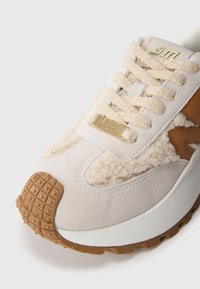 Sneaker bej cu un overlay texturat pufos, parte superioară din daim, talpă din cauciuc cu un grip pătat și un accent de logo auriu pe limbă.