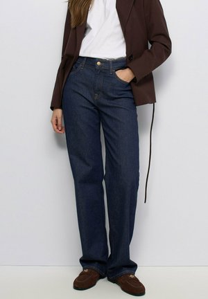 Mujer con vaqueros azul oscuro de pierna recta, camiseta blanca, blazer marrón y mocasines marrones, de pie frente a una pared lisa y clara.