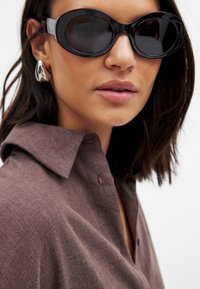 Mustad ovaalsed prillid, läikivad raamid. Pruun tekstuuriga särk kraega. Hõbedased silmapaistvad kõrvarõngad. Sile nahk, neutraalne huulevärv.