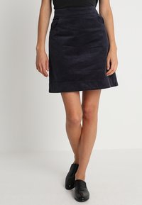Marineblauwe corduroyen mini-jurk met voorzakken, met een gestructureerd oppervlak en een rechte silhouet, gecombineerd met zwarte platte schoenen.
