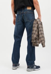 Vaqueros de mezclilla azul oscuro con diseño de pierna recta, que cuentan con bolsillos traseros y un parche de cuero con la marca. El modelo sostiene una chaqueta a cuadros.