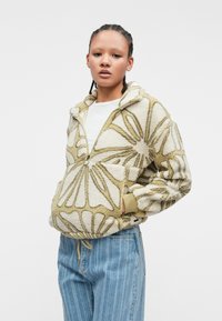 Fleece zip-up jakna v kremni in olivno zeleni barvi z cvetličnim vzorcem, sprednji žep in elastičnimi manšeti. Naročena preko bele srajce in modrih kavbojk.