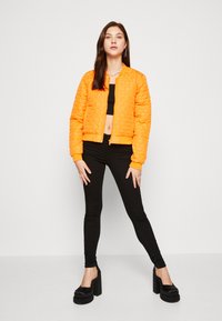 Chaqueta bomber acolchada naranja con puños y dobladillo de canalé, llevada sobre un top corto negro y combinada con leggings ajustados negros y zapatos de plataforma.