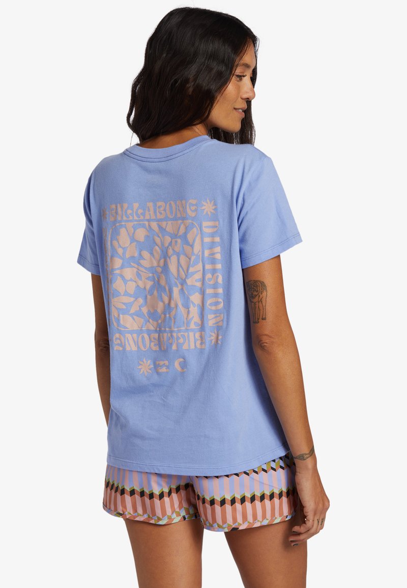 Camiseta de algodón azul claro con un gran estampado gráfico beige en la espalda que presenta un diseño floral y el nombre de la marca. Combinada con pantalones cortos de estampado.