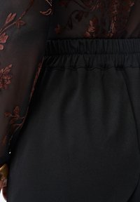 Close-up van een zwarte elastische tailleband van een broek met een doorschijnende zwarte mouw met bruine bloemrijke borduursels.