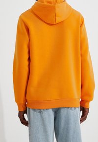 Sudadera naranja con bolsillo tipo canguro, puños y dobladillo acanalados; hecha de una suave mezcla de algodón. Llevada con jeans de mezclilla azul claro.