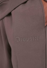 Nahaufnahme von mauvefarbenen Jogginghosen mit dem eingeprägten Schriftzug "Dropsize" nahe der Vordertasche, getragen von einer Person mit einer Hand in der Tasche.