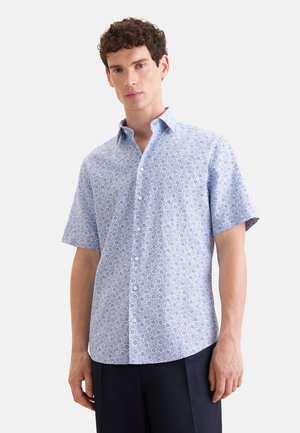Uomo che indossa una camicia azzurro chiaro a maniche corte con un piccolo motivo circolare, abbinata a pantaloni scuri, in piedi davanti a uno sfondo uniforme.