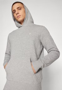 Ung mand iført lysegrå hættetrøje med lille logo på brystet, hænderne i foranlommen, kigger let opad mod en ensfarvet baggrund.