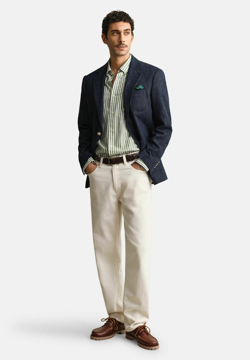 Uomo in piedi con le mani in tasca, indossa una giacca blu navy, una camicia a righe verdi, pantaloni color crema e scarpe di pelle marrone su uno sfondo bianco.